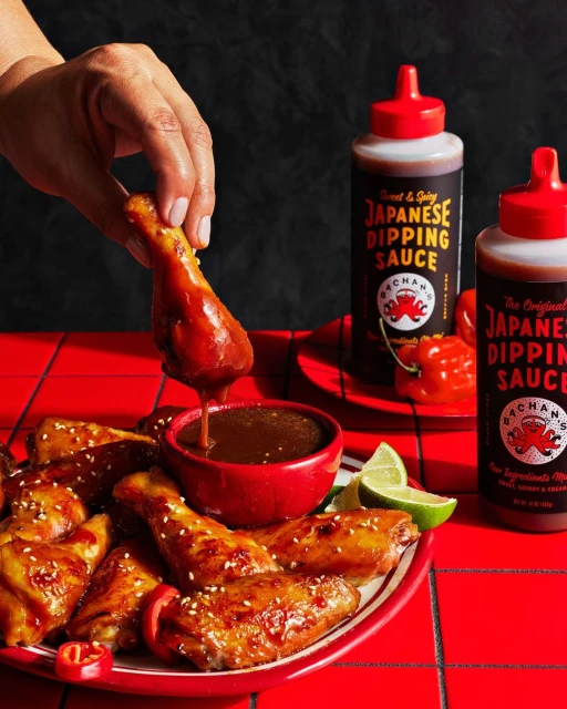 A hand dips a fried chicken wing into a red bowl of dipping sauce alongside a platter of chicken wings, limes, and sauce bottles.
