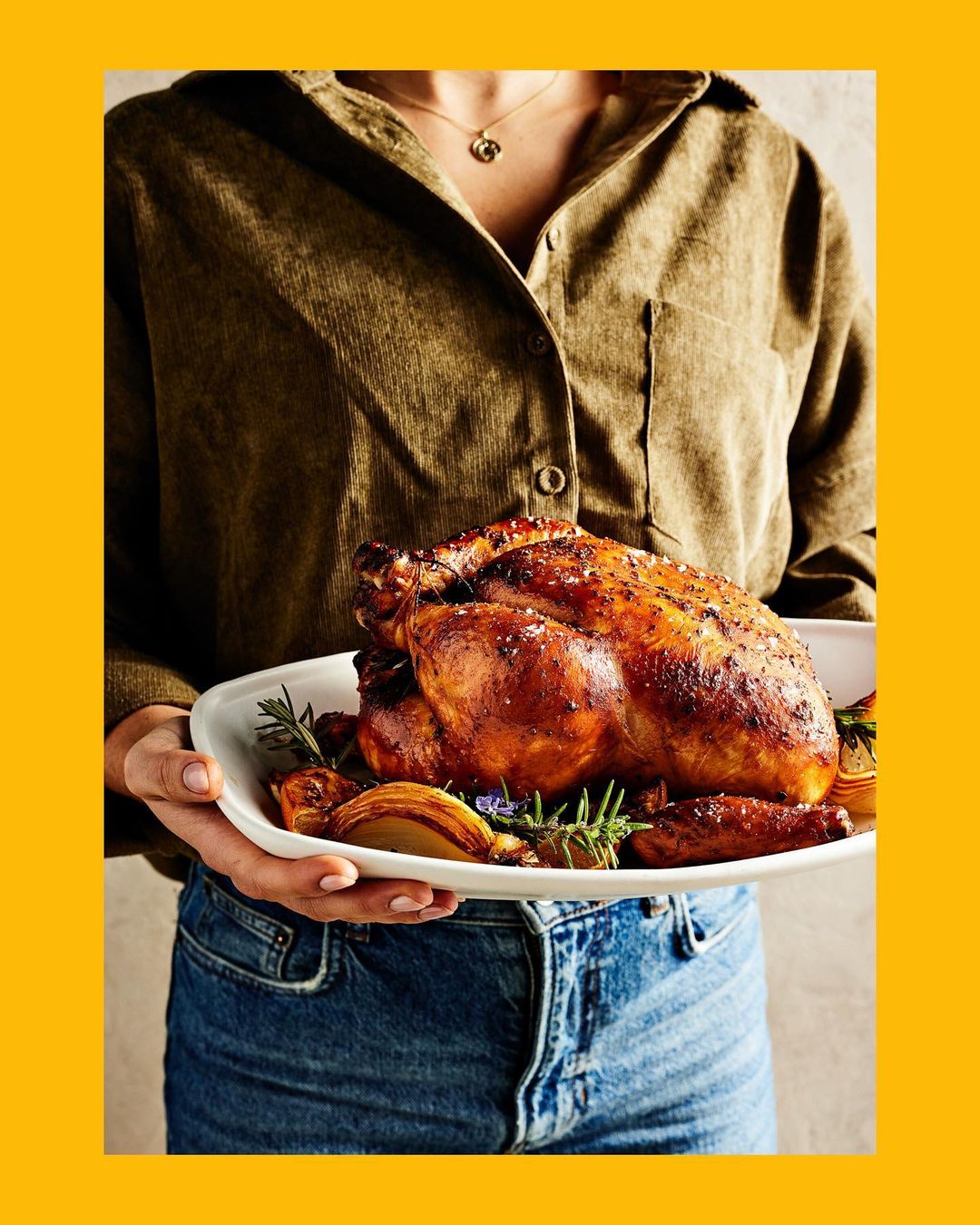 Roasted chicken on a white platter held by a person in a corduroy shirt and jeans against a yellow background. Food photography.