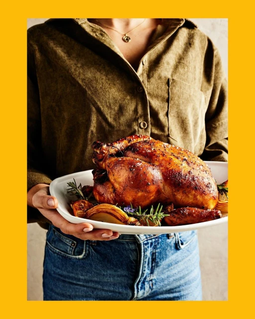 Roasted chicken on a white platter held by a person in a corduroy shirt and jeans against a yellow background. Food photography.