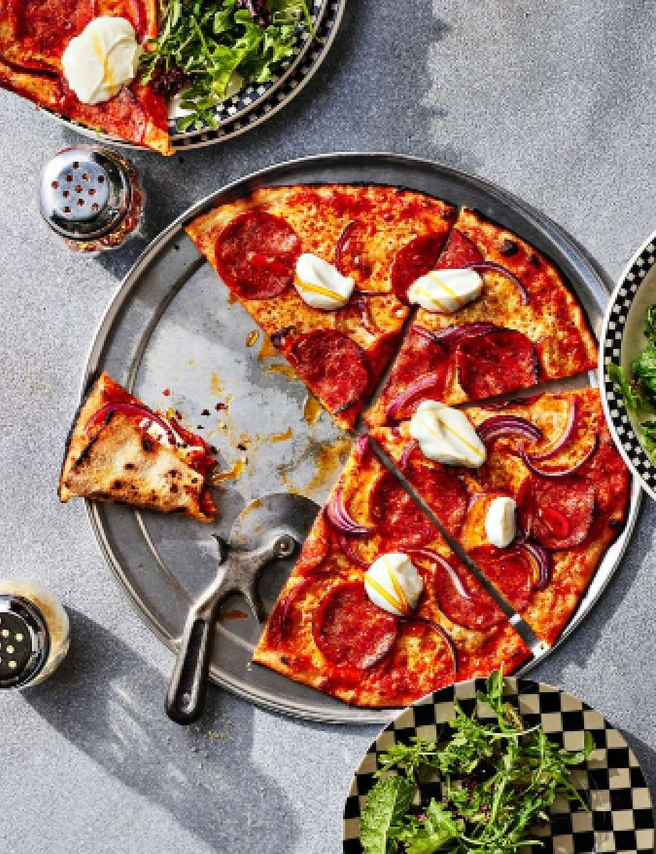 Overhead shot of a sliced pepperoni pizza with dabs of cheese and red onion on a metal tray, with salad and salt shakers around it.
