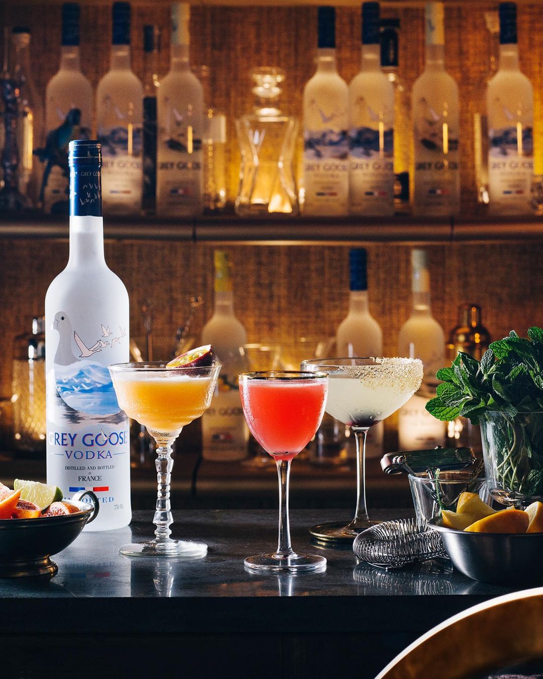 Three colorful cocktails in stemmed glasses, positioned in front of a Grey Goose vodka bottle and a bar shelf stocked with bottles.