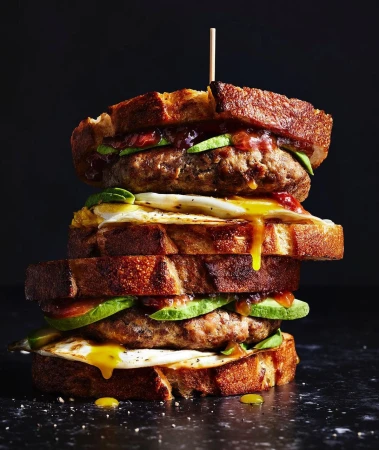 Towering sandwich with toasted bread, egg, meat, and avocado filling layers, set against a dark backdrop; ideal for food advertising.