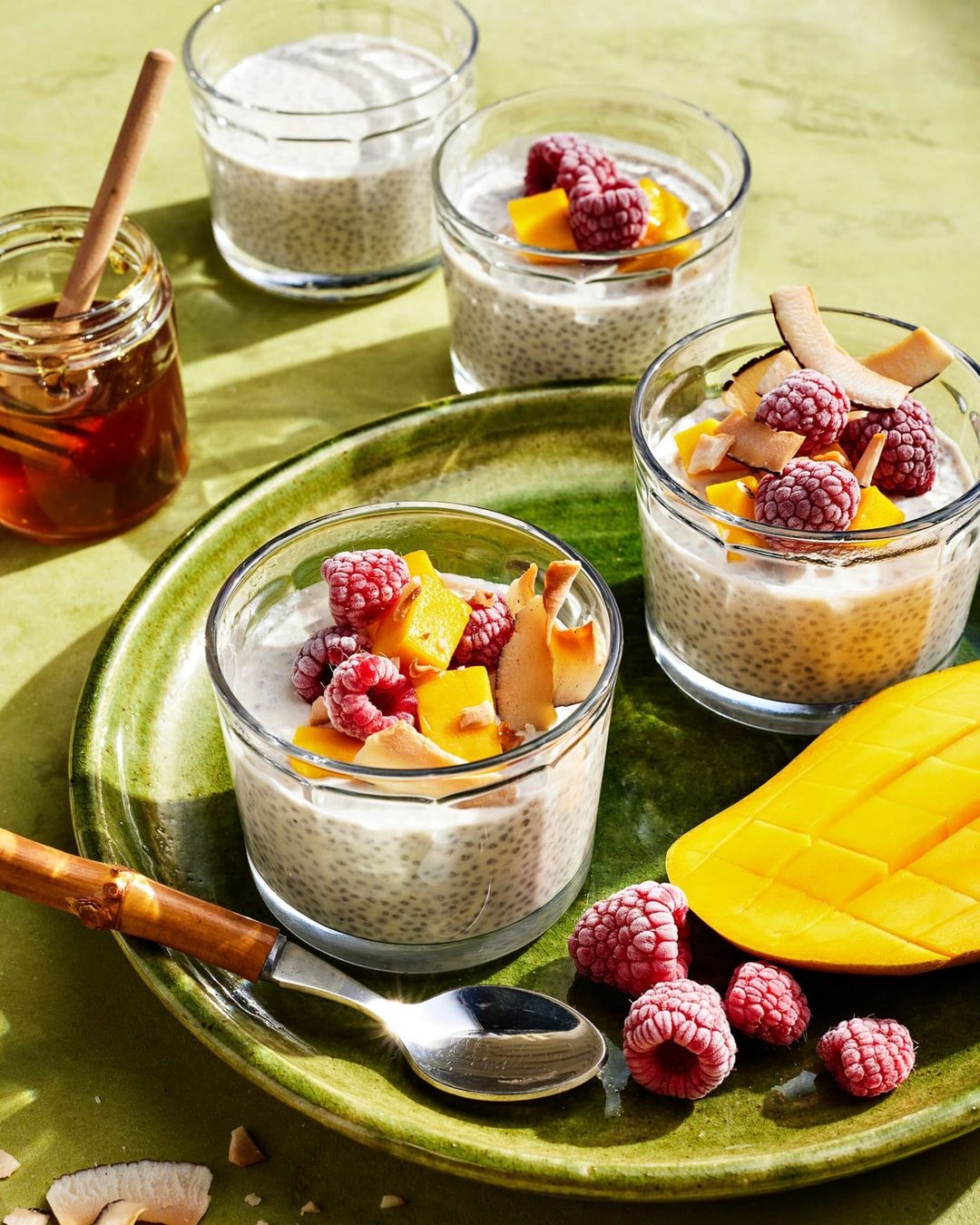Chia seed puddings in clear glass jars topped with raspberries, mango, and coconut flakes, served with honey and a silver spoon.