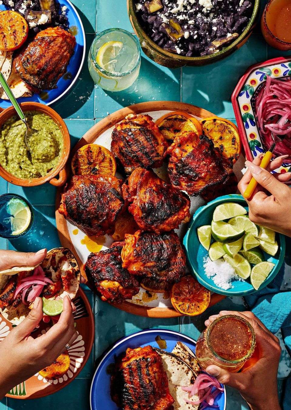 Overhead shot of a vibrant feast featuring grilled chicken thighs, citrus slices, salsa, tacos, and a variety of colorful accompaniments on a tile surface.
