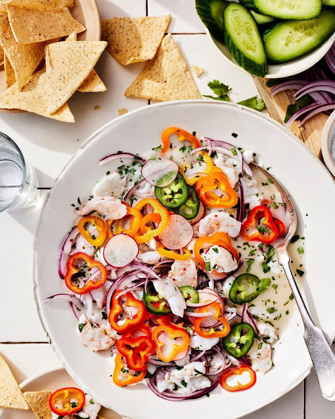 Overhead shot of shrimp ceviche with red onion, jalapenos, radish, and sweet peppers in a white bowl with tortilla chips on the side.