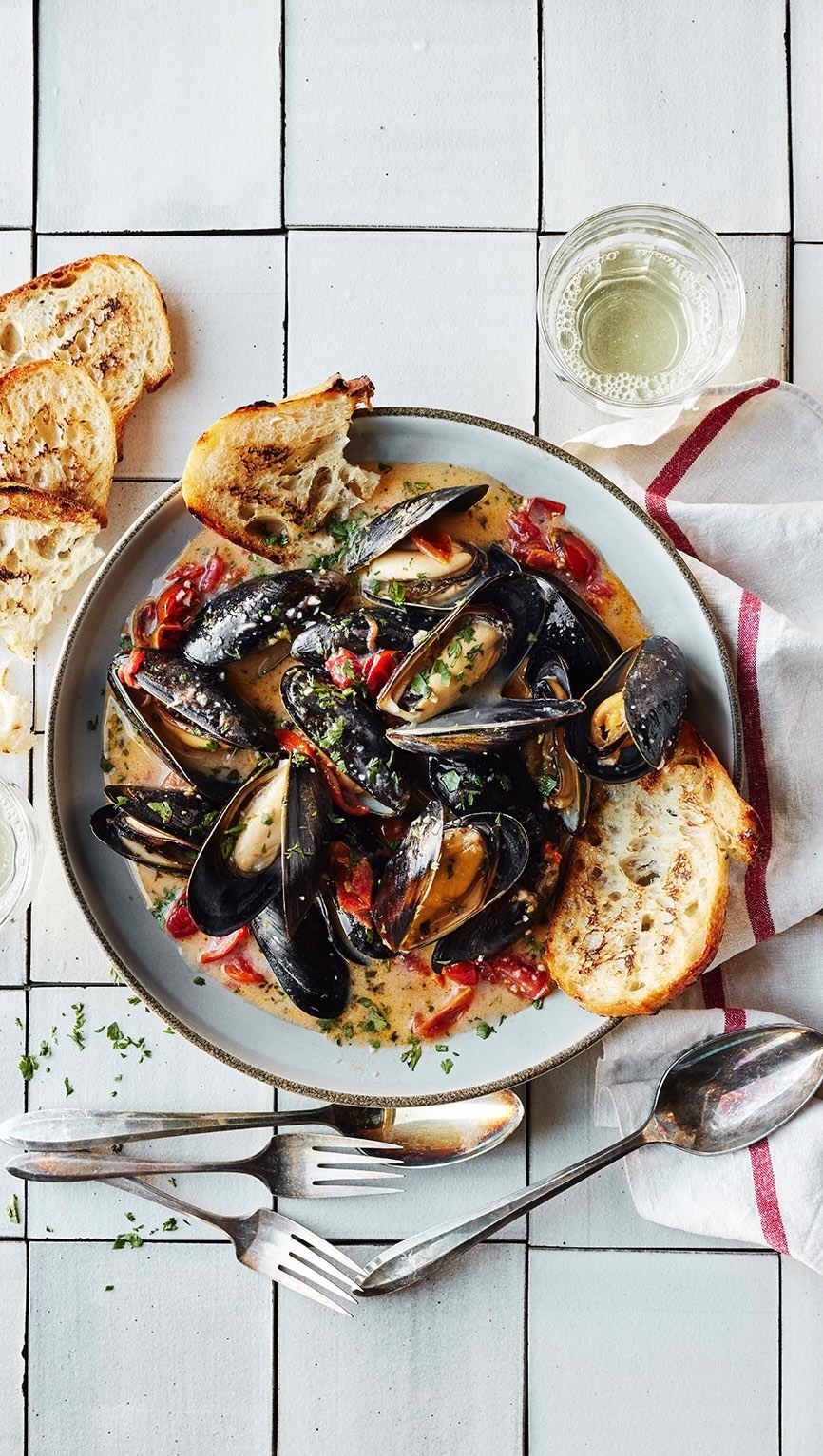 Overhead shot of a ceramic plate filled with steamed mussels, garnished with herbs and accompanied by grilled bread and silverware.