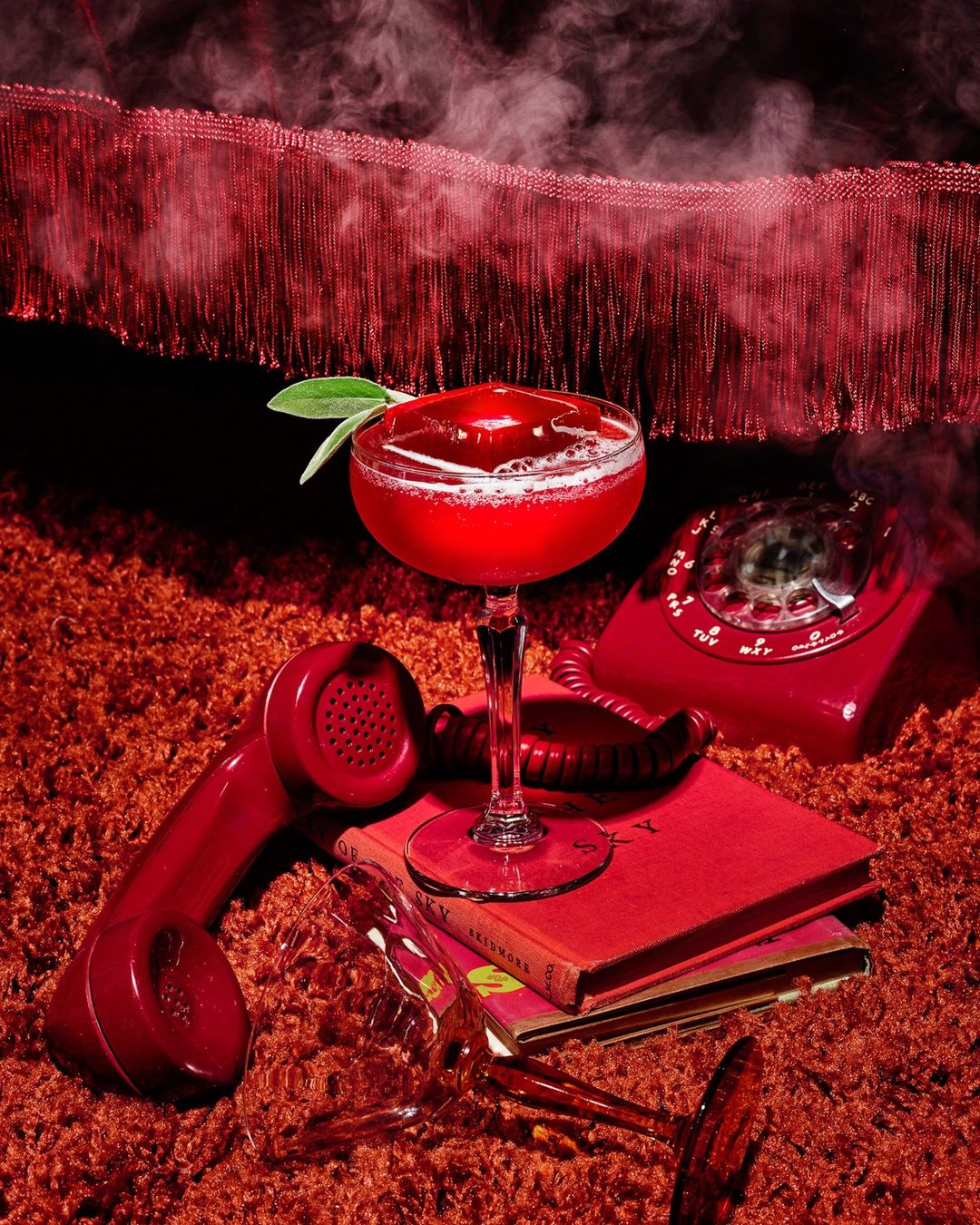 Red-themed still life: a cocktail in a coupe glass, vintage rotary phone, and books are artfully arranged on a shag carpet.