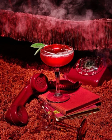 Red-themed still life: a cocktail in a coupe glass, vintage rotary phone, and books are artfully arranged on a shag carpet.