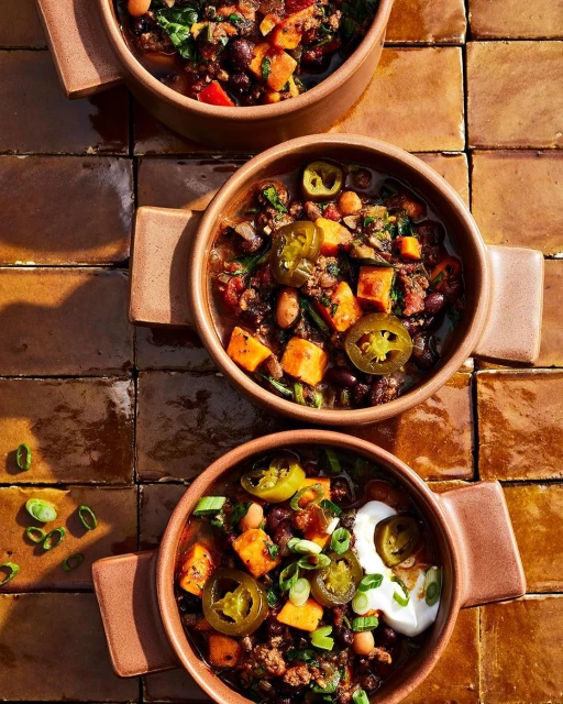 Overhead shot of three bowls of chili with sweet potatoes, beans, jalapenos, and sour cream on a brown tiled surface. Food photography.