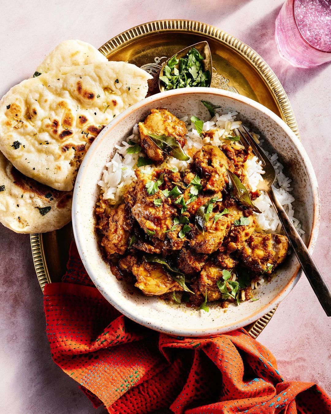 Overhead shot of a bowl of chicken curry and rice, accompanied by naan bread and a glass of pink liquid, on a brass tray.
