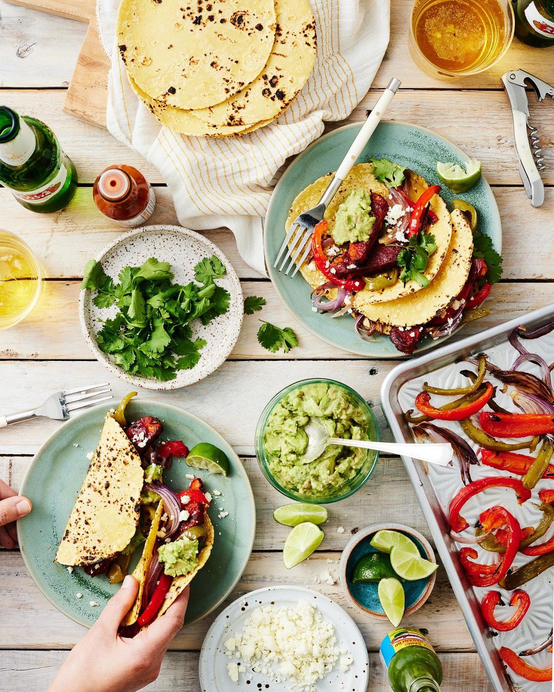 Overhead shot of tacos on plates with various toppings, limes, cilantro, and beer, arranged on a white wooden table.
