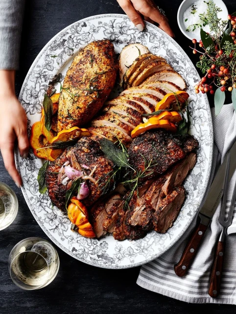 Overhead shot of a floral-patterned platter filled with carved and cooked meats, squash, and herbs, placed on a grey stone surface.