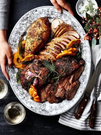 Overhead shot of a floral-patterned platter filled with carved and cooked meats, squash, and herbs, placed on a grey stone surface.