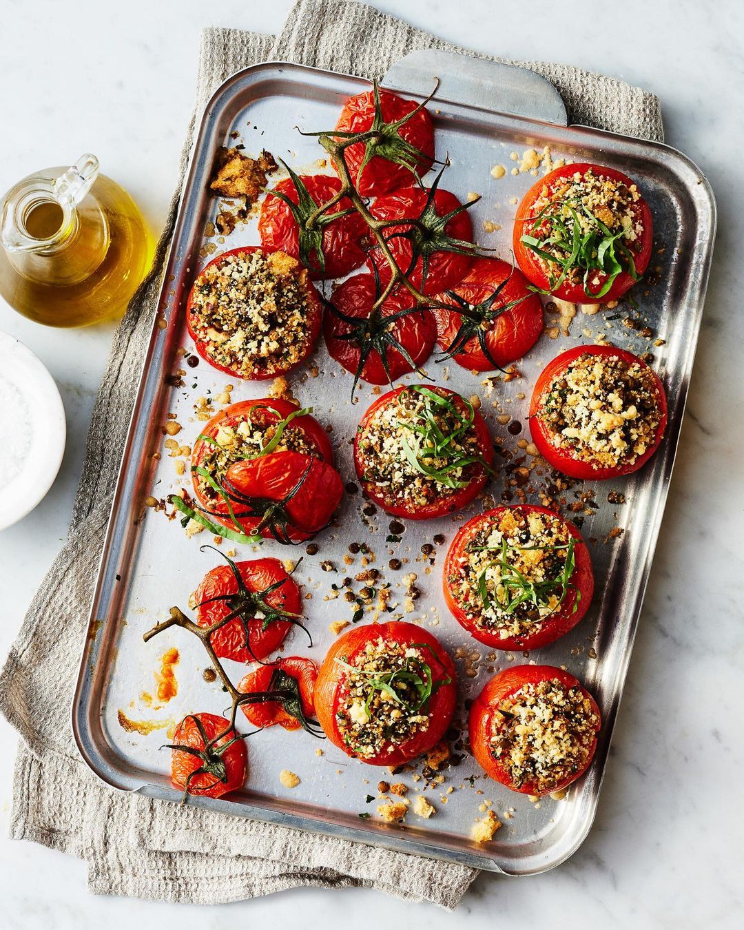 Overhead shot of baked tomatoes stuffed with breadcrumbs and greens, arranged on a silver tray, with olive oil and salt.