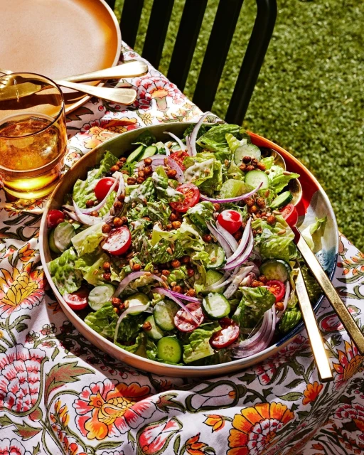 Overhead view of a colorful salad with lettuce, cucumbers, tomatoes, onions, and chickpeas on a floral tablecloth outdoors.
