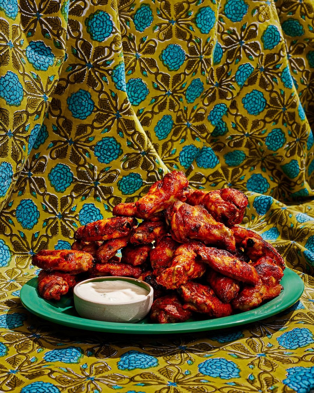 Pile of spicy chicken wings on a green plate with a bowl of dipping sauce against a patterned yellow and turquoise fabric backdrop.