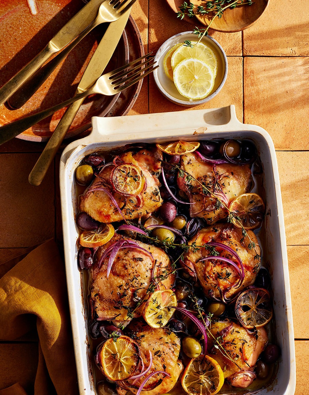 Overhead shot of chicken thighs with lemon slices, olives, and red onions in a baking dish, accompanied by cutlery and dishware.