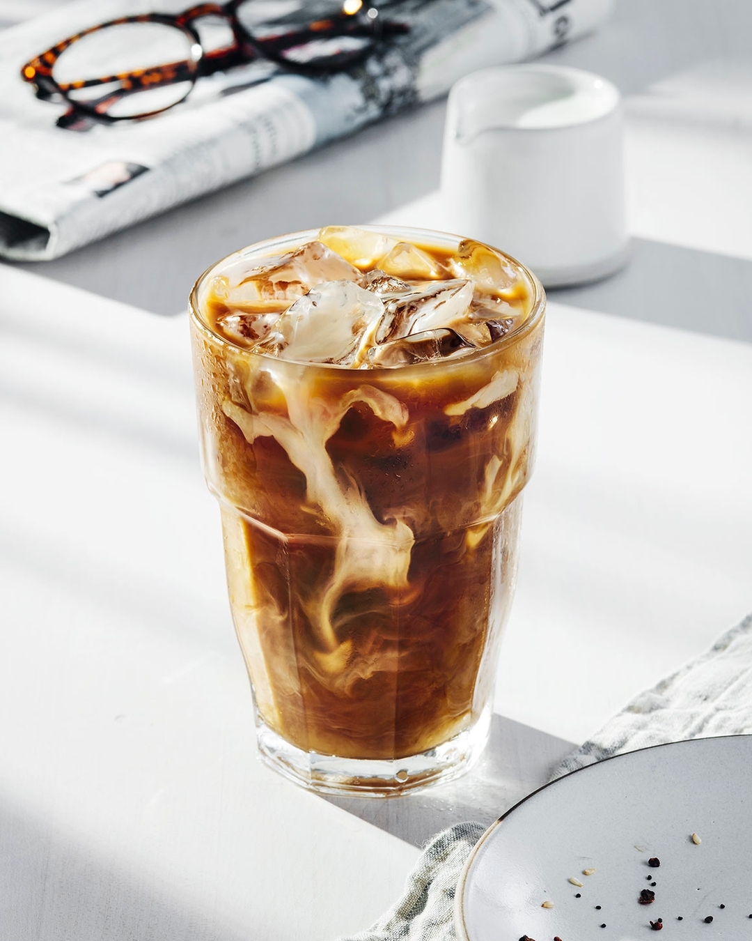 Iced coffee drink with milk in a glass on a white table, with a plate, newspaper, and glasses in the background, bright light.