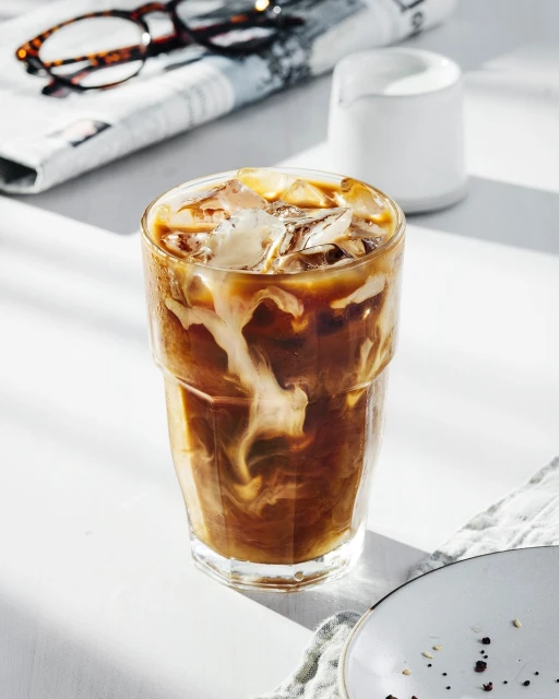 Iced coffee drink with milk in a glass on a white table, with a plate, newspaper, and glasses in the background, bright light.