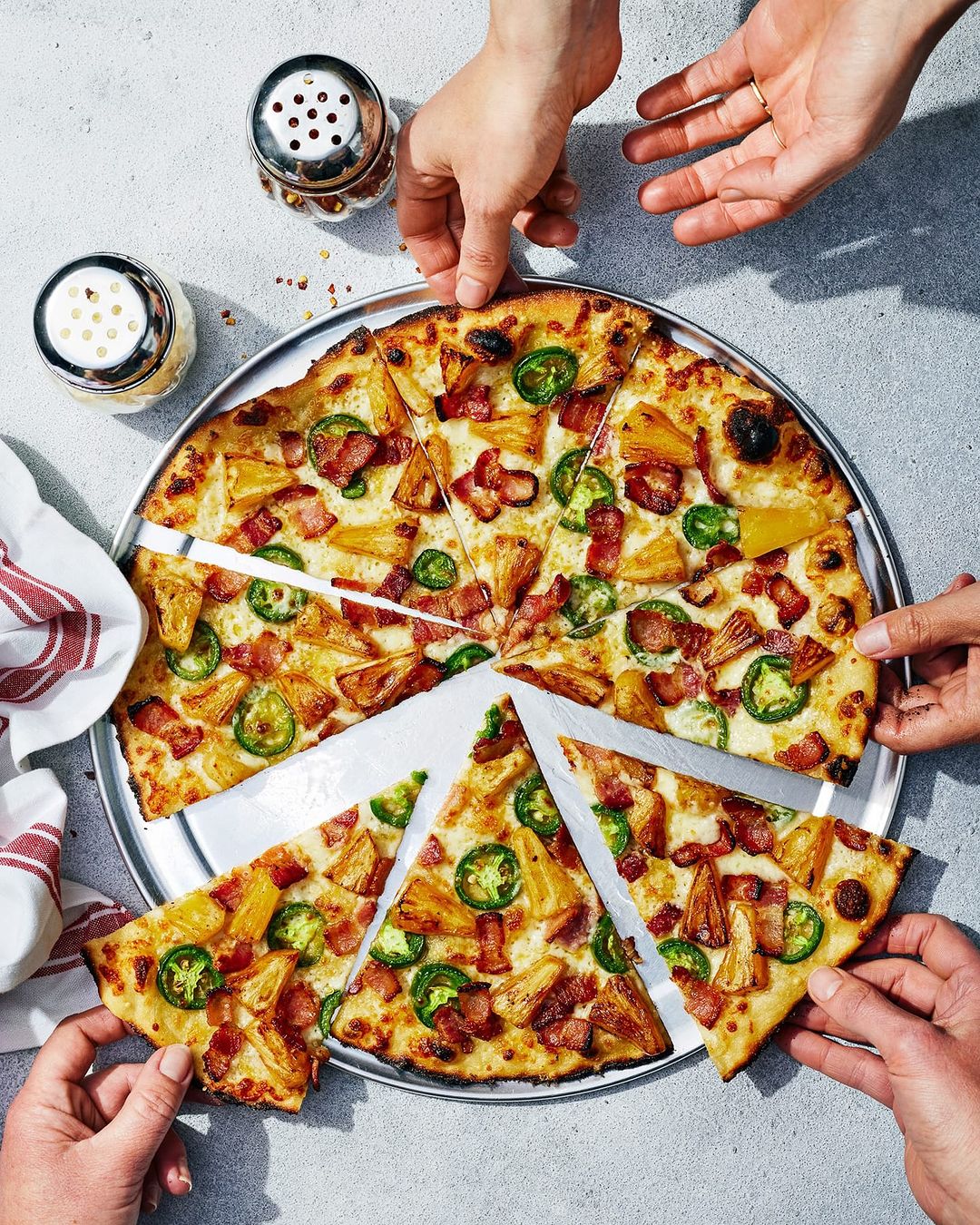 Overhead shot of a sliced pizza on a metal tray, featuring pineapple, bacon, and jalapeños, with several hands reaching for slices.