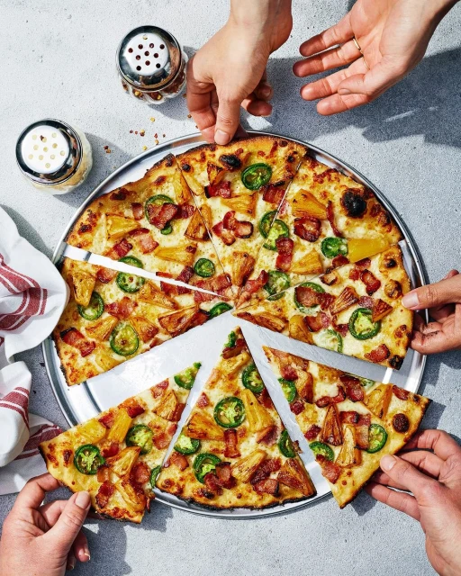 Overhead shot of a sliced pizza on a metal tray, featuring pineapple, bacon, and jalapeños, with several hands reaching for slices.