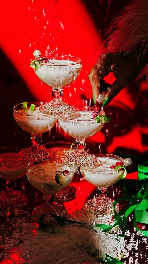 Tower of coupe glasses filled with white cocktails, garnished with cherries and lime peels, being dusted with white powder against red background.