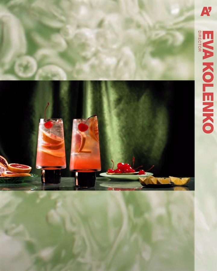 Two highball cocktails with grapefruit, cherry garnish, and ice cubes sit on a reflective surface against a dark green background.