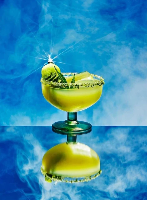 Brightly lit glass with a green cocktail garnished with a lime wedge sits atop a reflective surface with a cloudy blue background.