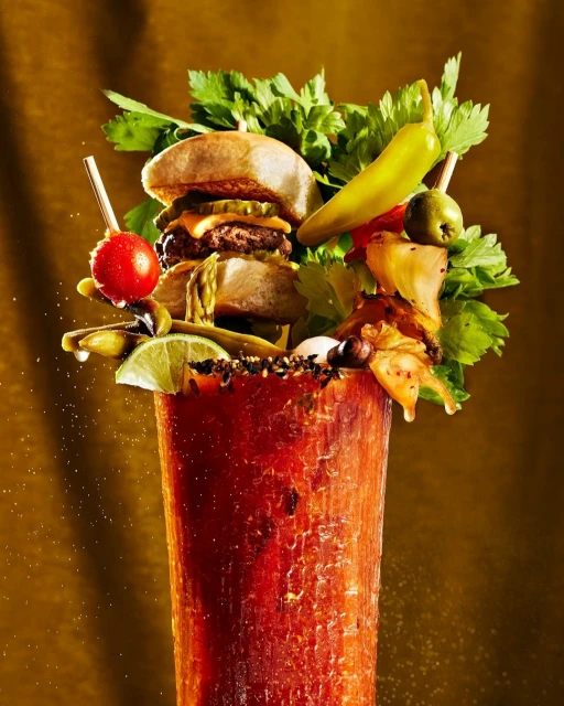 Tall cocktail glass filled with a bloody mary and garnished with a miniature burger, celery, tomatoes, lime wedge, and pepperoncini.
