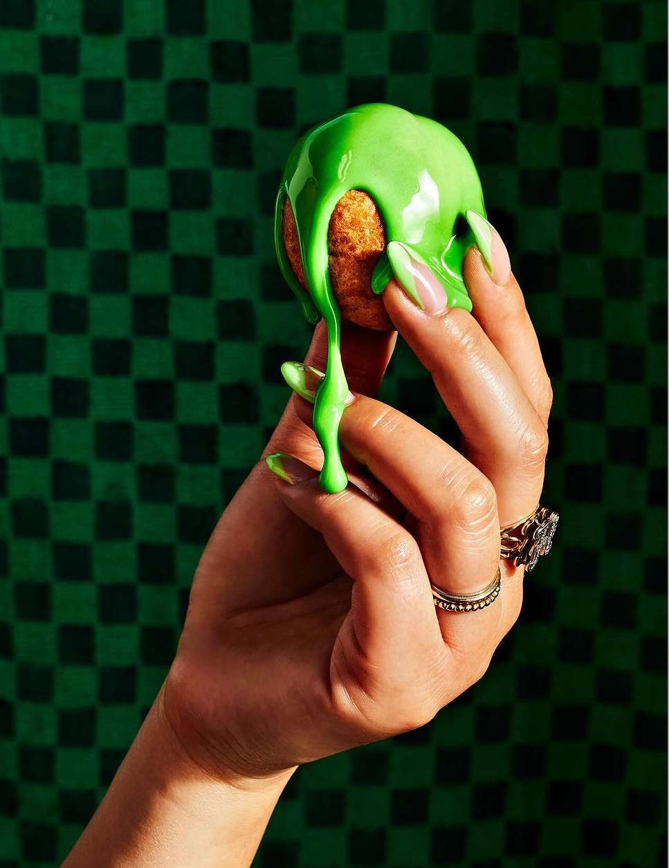 Close-up shot of a tan baked good covered in bright green glaze, held by a light-skinned hand with multiple rings and green painted nails.