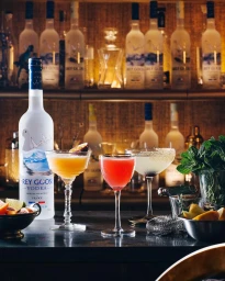 Cocktail bar display featuring a vodka bottle and three colorful cocktails on a dark counter against a backlit shelving unit.