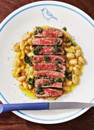 Sliced grilled steak with green herb chimichurri sauce served over white beans on a white plate with a blue-handled knife.