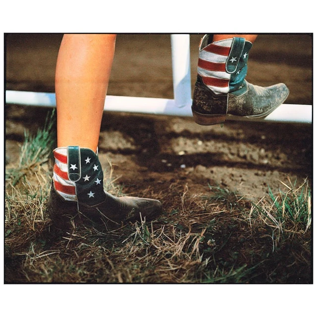Cowboy boots with US flag design standing on a white rail. Outdoor scene with grass and dirt.