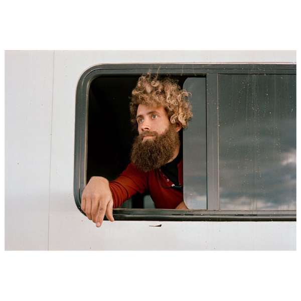 Man with beard and curly hair looks out of a vehicle window. He is wearing a red shirt and has one hand visible.