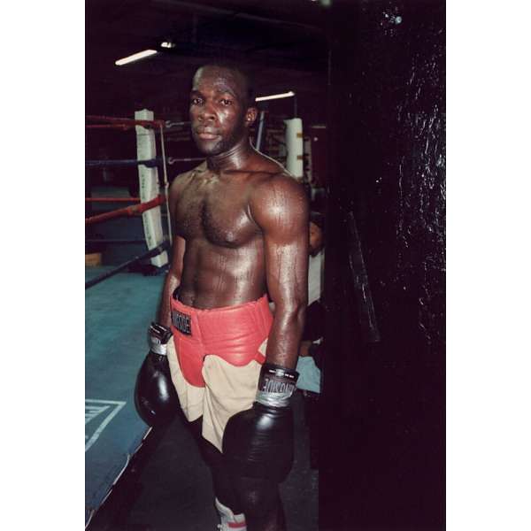 Boxer in red shorts and black gloves stands in a boxing gym near a ring, looking serious and athletic. 
