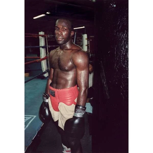 Boxer in red shorts and black gloves stands in a boxing gym near a ring, looking serious and athletic. 
