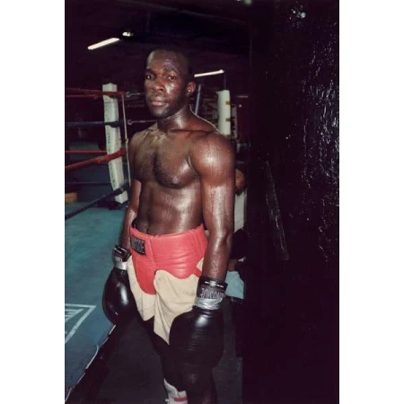Boxer in red shorts and black gloves stands in a boxing gym near a ring, looking serious and athletic. 