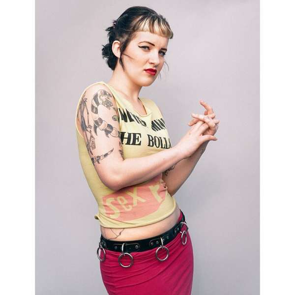 A young woman with fair skin, wearing a graphic tank top and pink skirt, poses against a neutral background in a studio shot.
