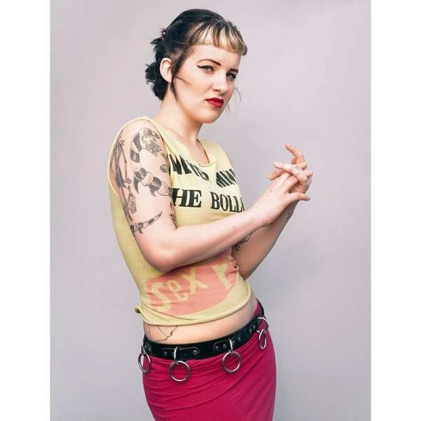 A young woman with fair skin, wearing a graphic tank top and pink skirt, poses against a neutral background in a studio shot.