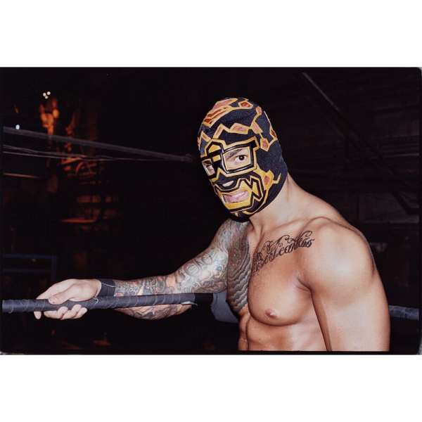 A muscular, bare-chested wrestler with arm tattoos is shown wearing a black and gold mask, standing behind the wrestling ring's ropes.