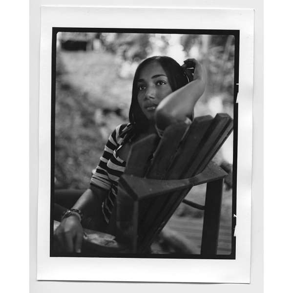 Monochrome portrait of a Black woman in a striped shirt sitting in a wooden Adirondack chair outside with blurred foliage background.
