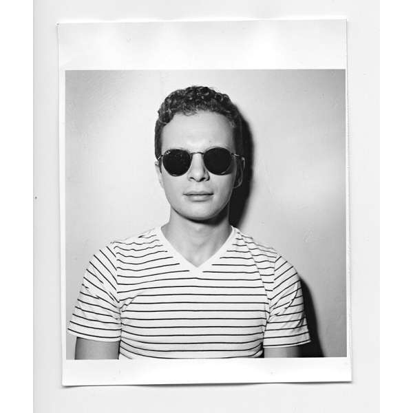 A medium shot of a young adult man wearing sunglasses and a striped shirt, set against a neutral indoor background in black and white.