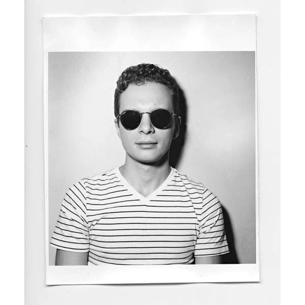 A medium shot of a young adult man wearing sunglasses and a striped shirt, set against a neutral indoor background in black and white.