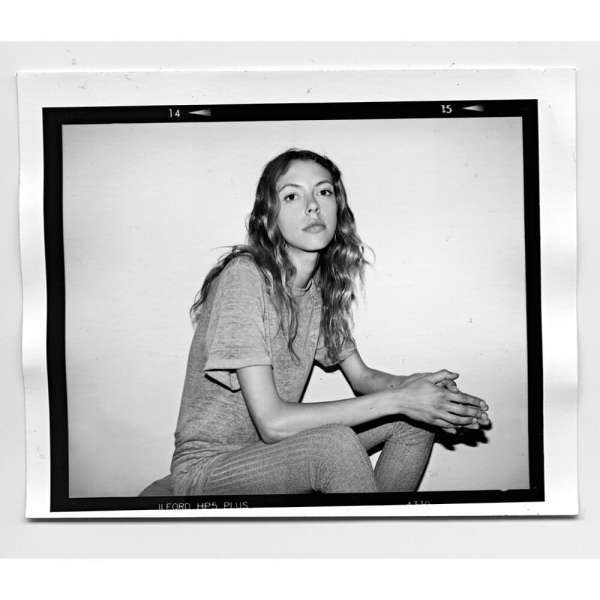 Black-and-white portrait of a young woman sitting indoors, wearing a grey t-shirt and leggings. She has long hair and a neutral expression.