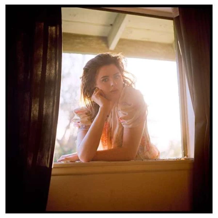 Woman leaning on a window sill looking out, framed by dark curtains with soft backlighting by Evan Klanfer.