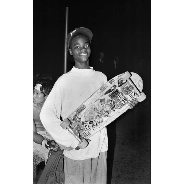 Monochrome photo of an African-American teenage boy with a cap, holding a skateboard with stickers. Another person is visible nearby.