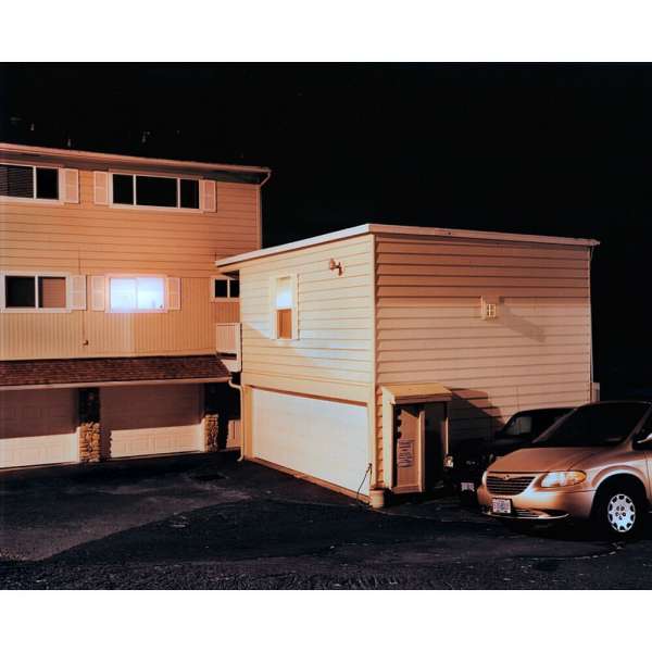 Exterior nighttime view of a multi-level tan house with a car parked in front, illuminated by warm artificial light from windows.