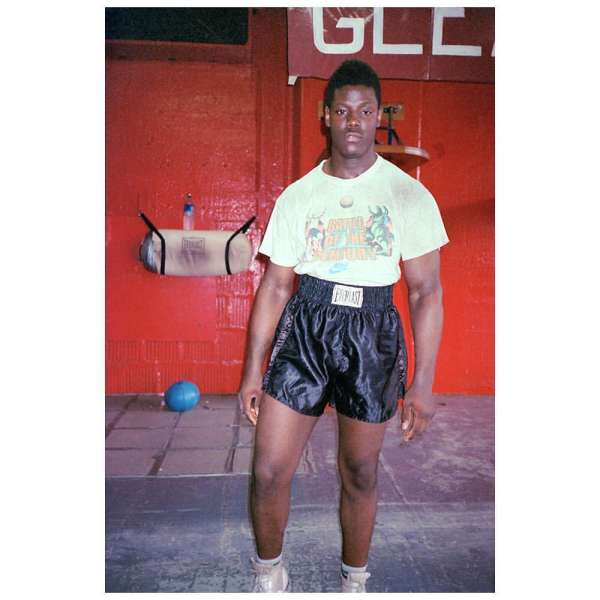 Muscular Black male boxer stands in a boxing gym wearing Everlast shorts, T-shirt, and sneakers near a hanging punching bag and blue medicine ball.