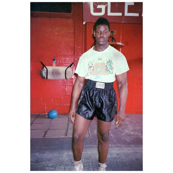 Muscular Black male boxer stands in a boxing gym wearing Everlast shorts, T-shirt, and sneakers near a hanging punching bag and blue medicine ball.