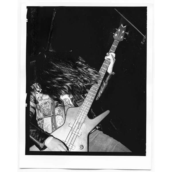 Black and white photo of a musician playing an electric bass guitar, wearing a jacket with patches on a dark stage.